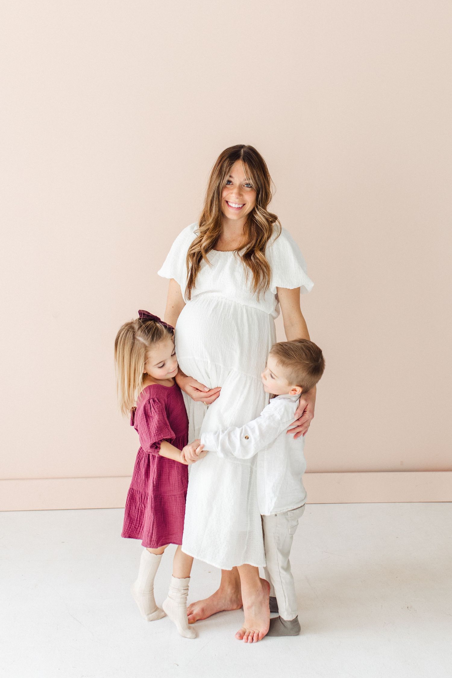 Family maternity photo with two children hugging pregnant mother in white dress.