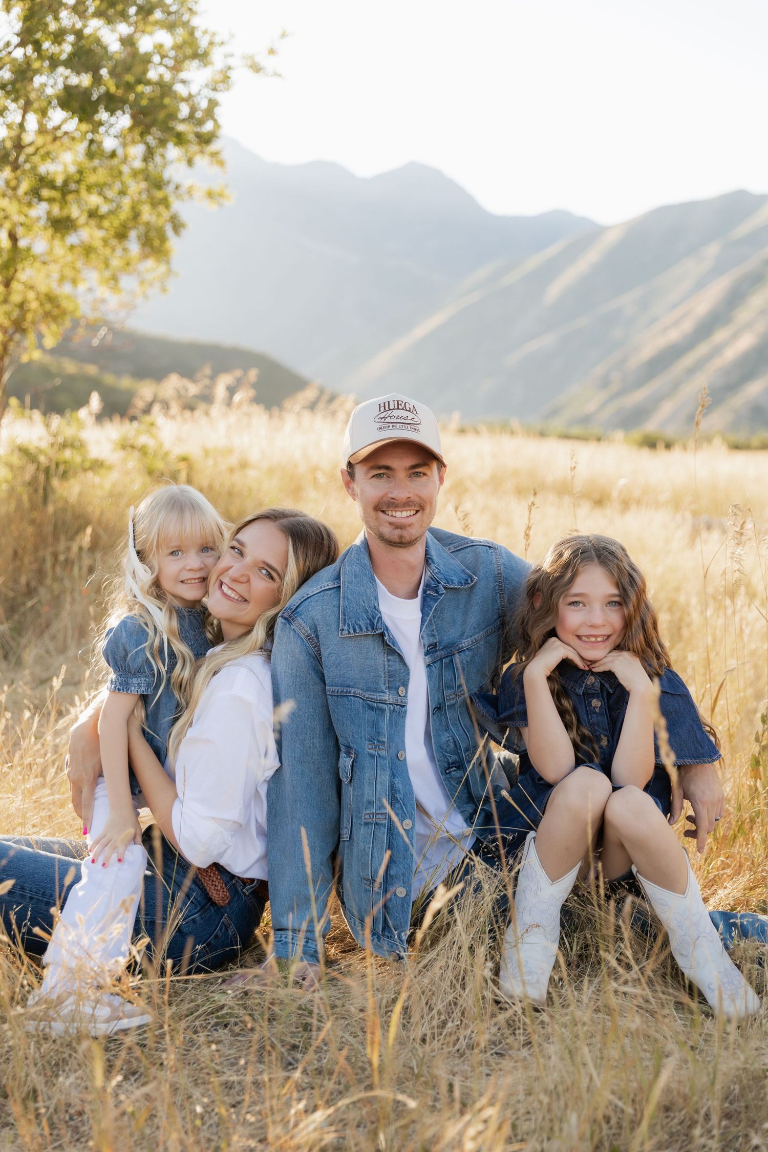 A family sits together in a golden field of tall grass with mountains in the background during sunset.