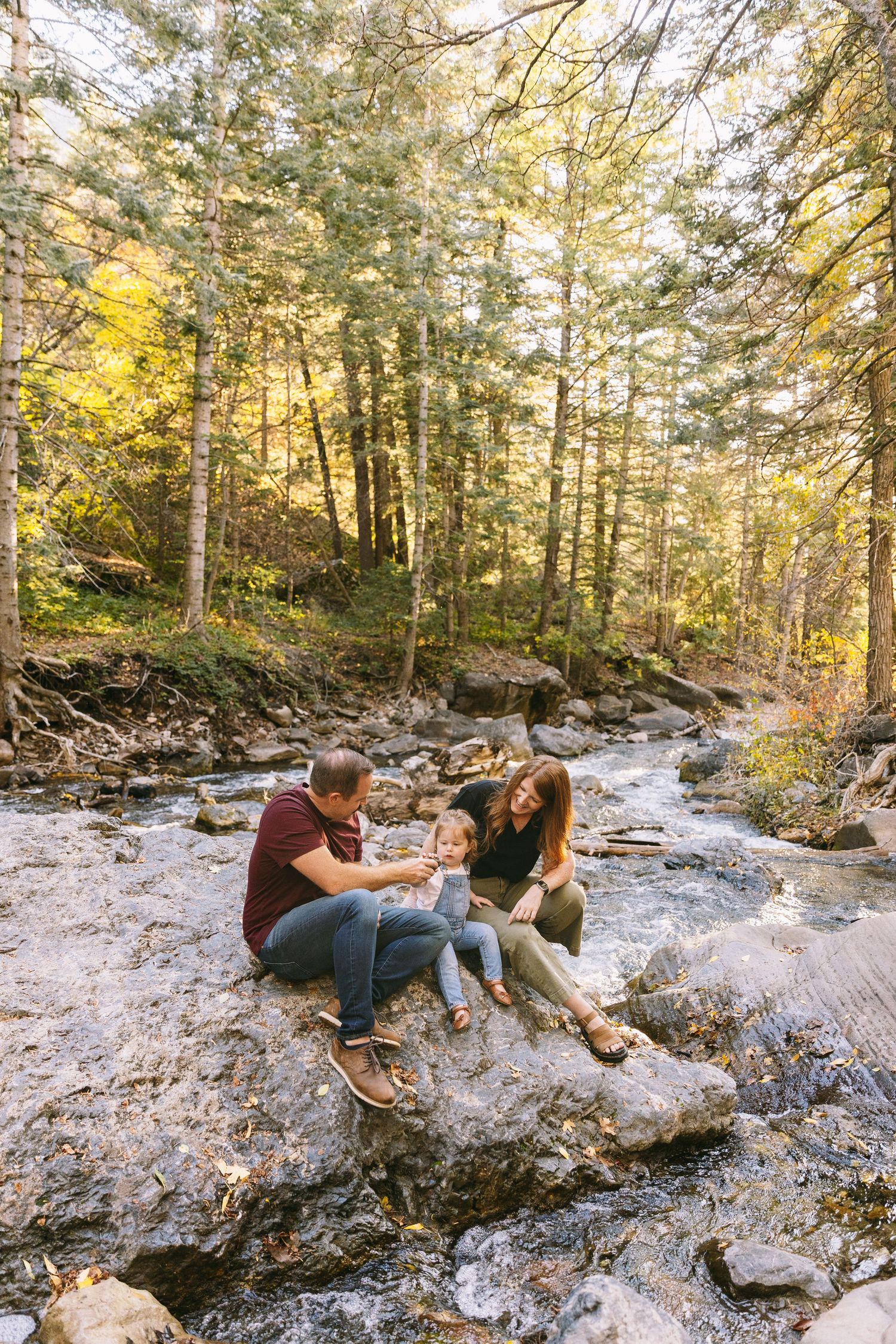 A couple relaxes on rocky terrain in a sunlit forest during an outdoor photo session.