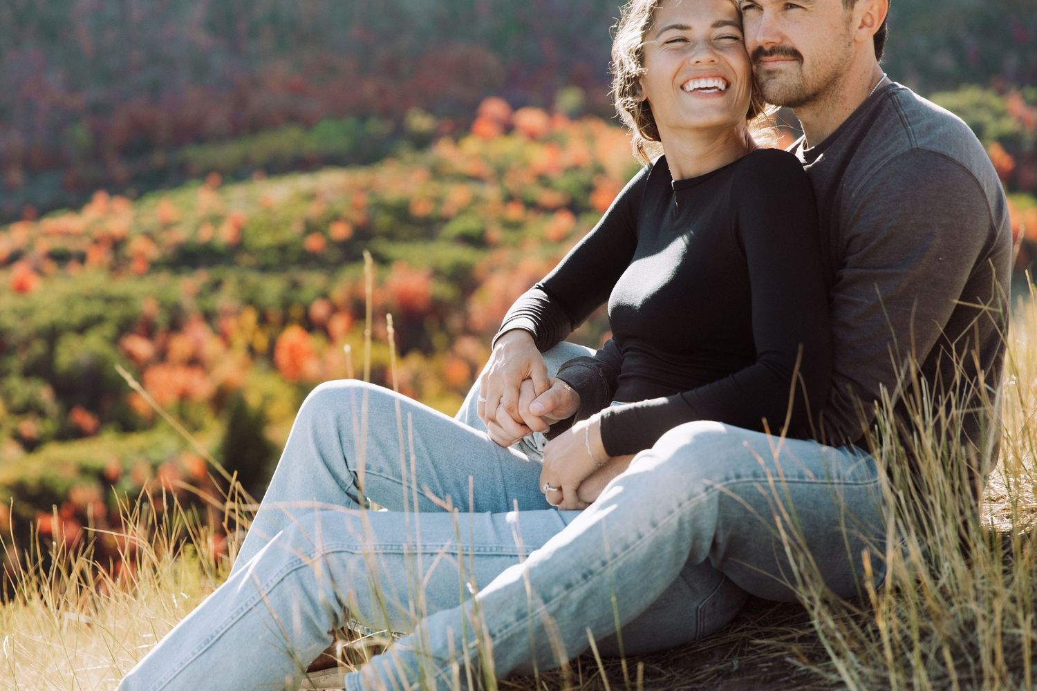 A couple shares a tender moment while sitting on a grassy hillside with autumn colors in the background.