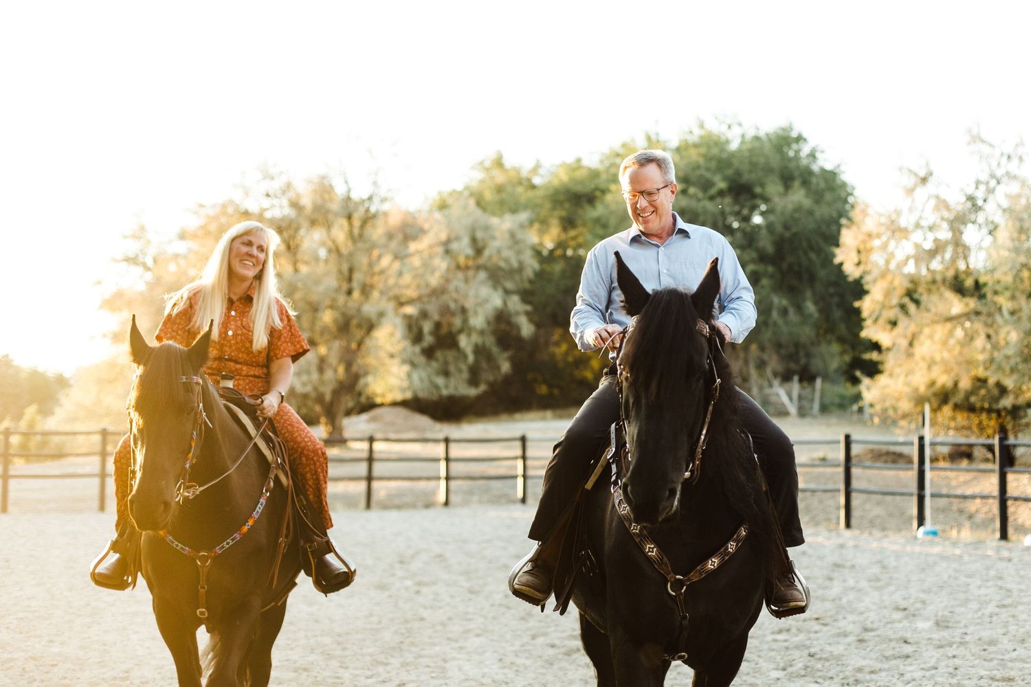 Two people riding black horses side by side in a paddock during golden hour.