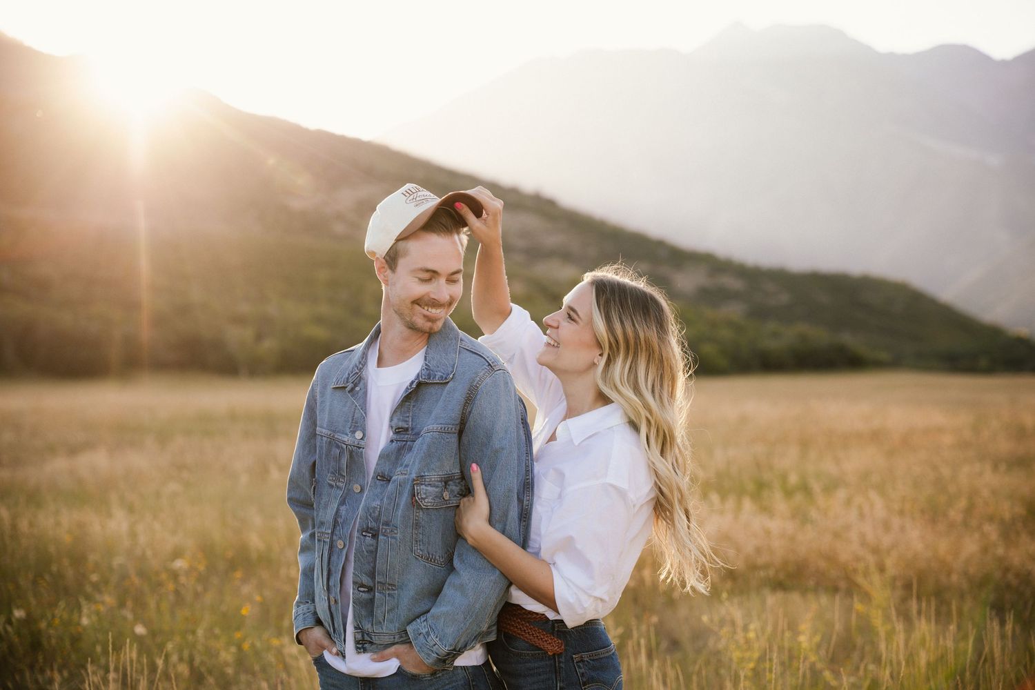 A couple embraces in a golden field at sunset with mountains in the background.