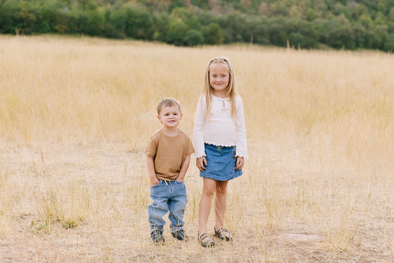 Two children stand together in a golden field of tall grass on a sunny day.