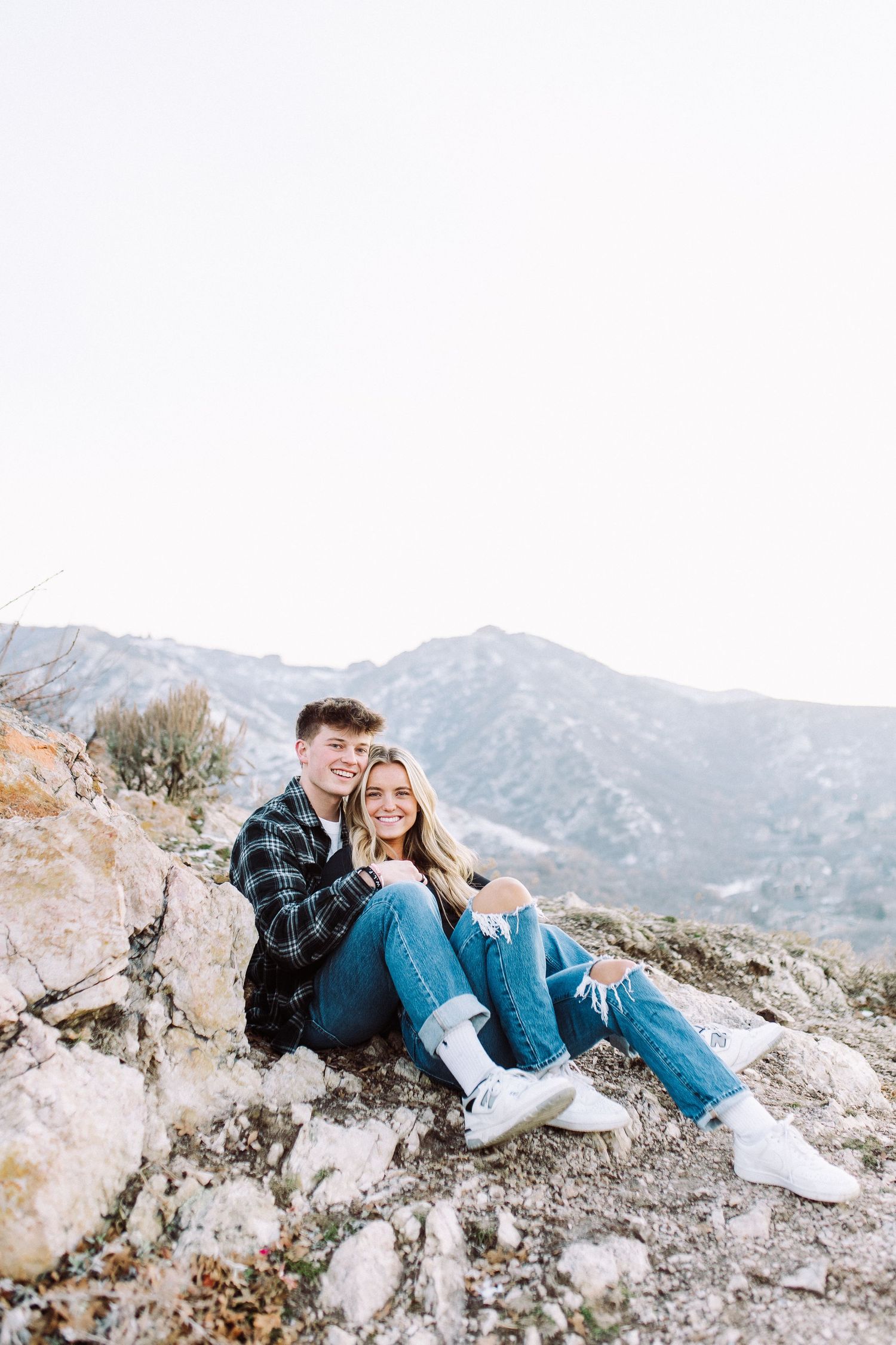 Couple in casual attire sitting on rocky mountain overlook with scenic mountain backdrop.
