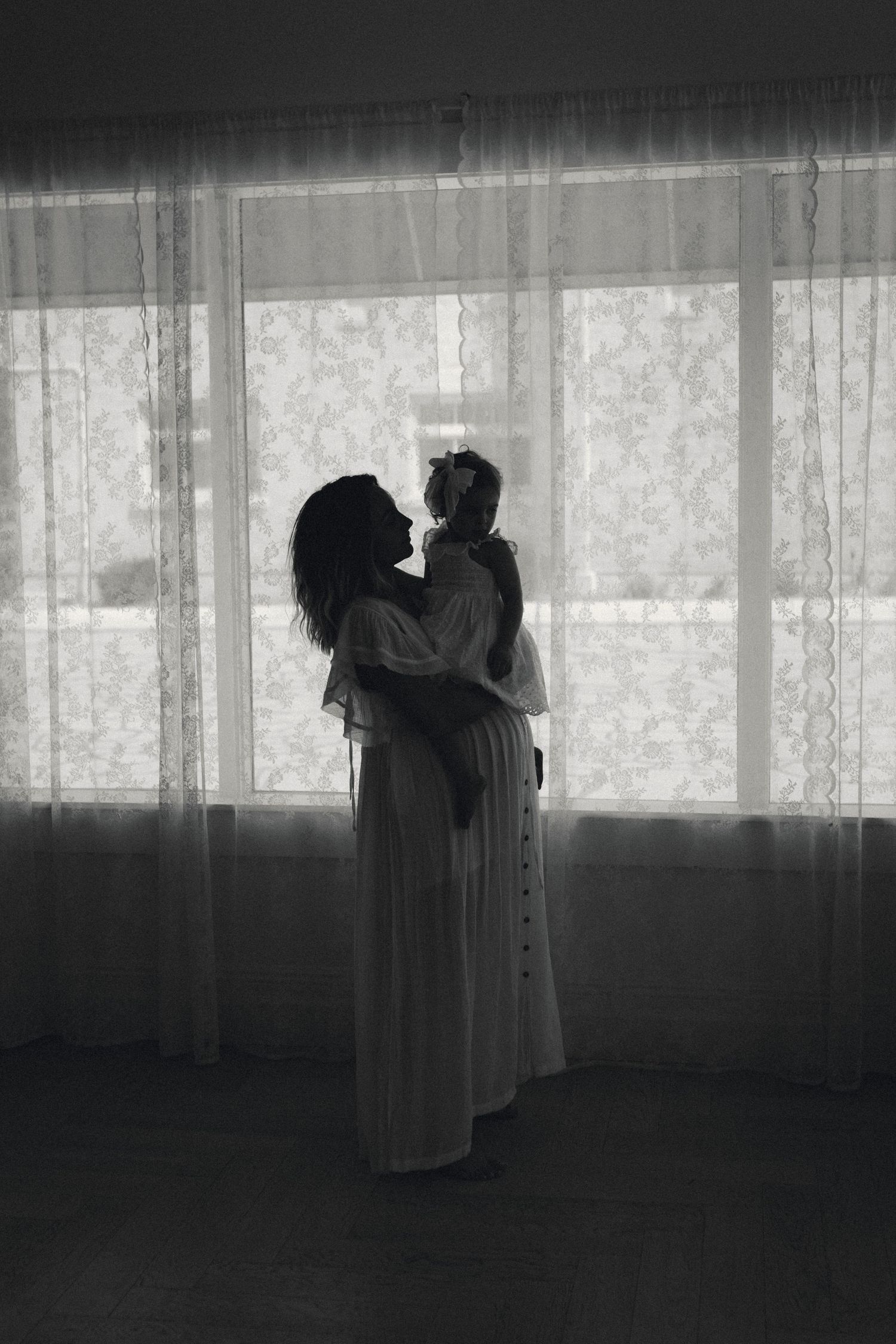 Silhouette of mother and child against backlit window with sheer curtains.