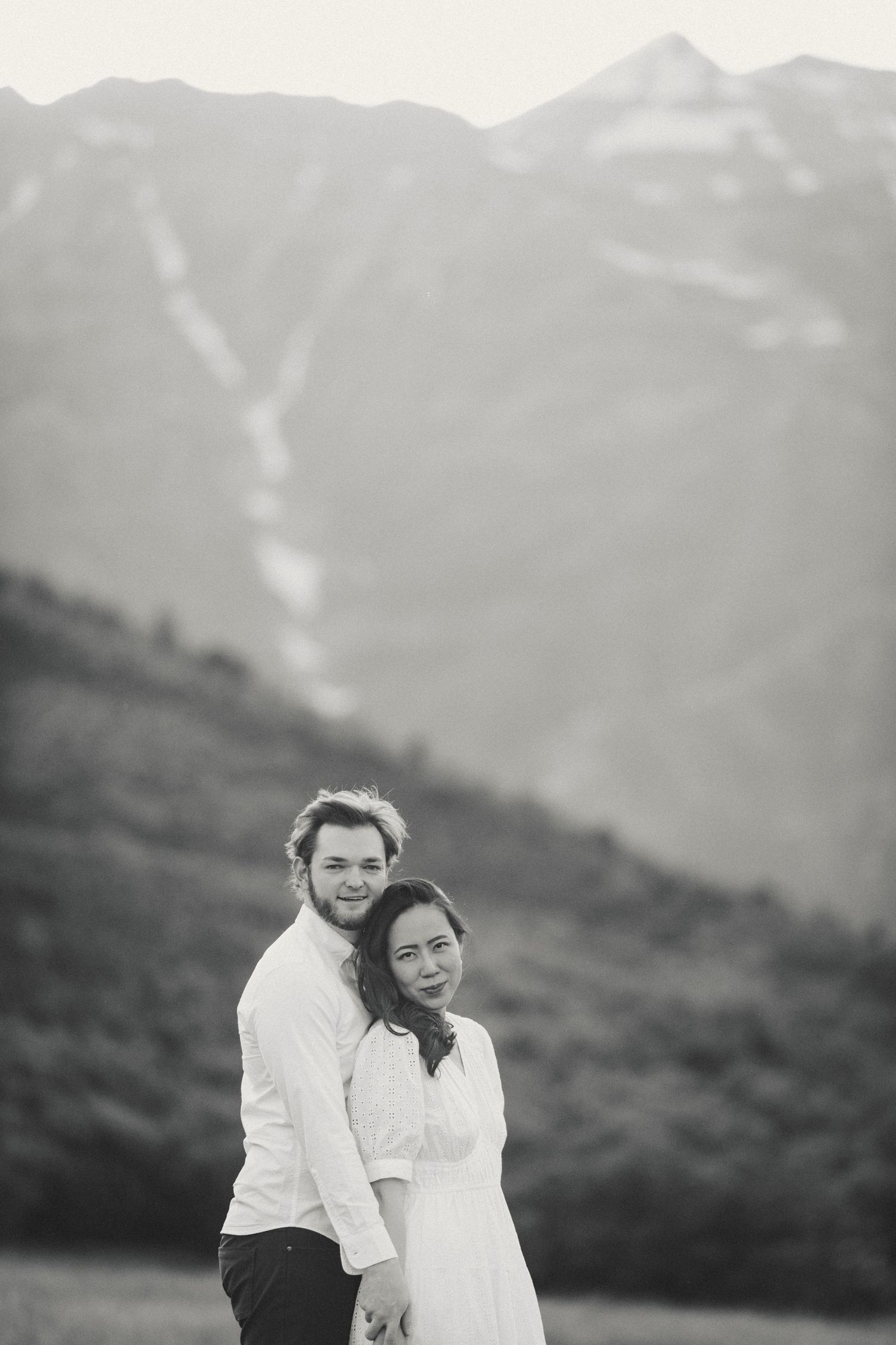 A couple in white outfits poses together against a scenic mountain backdrop in black and white.