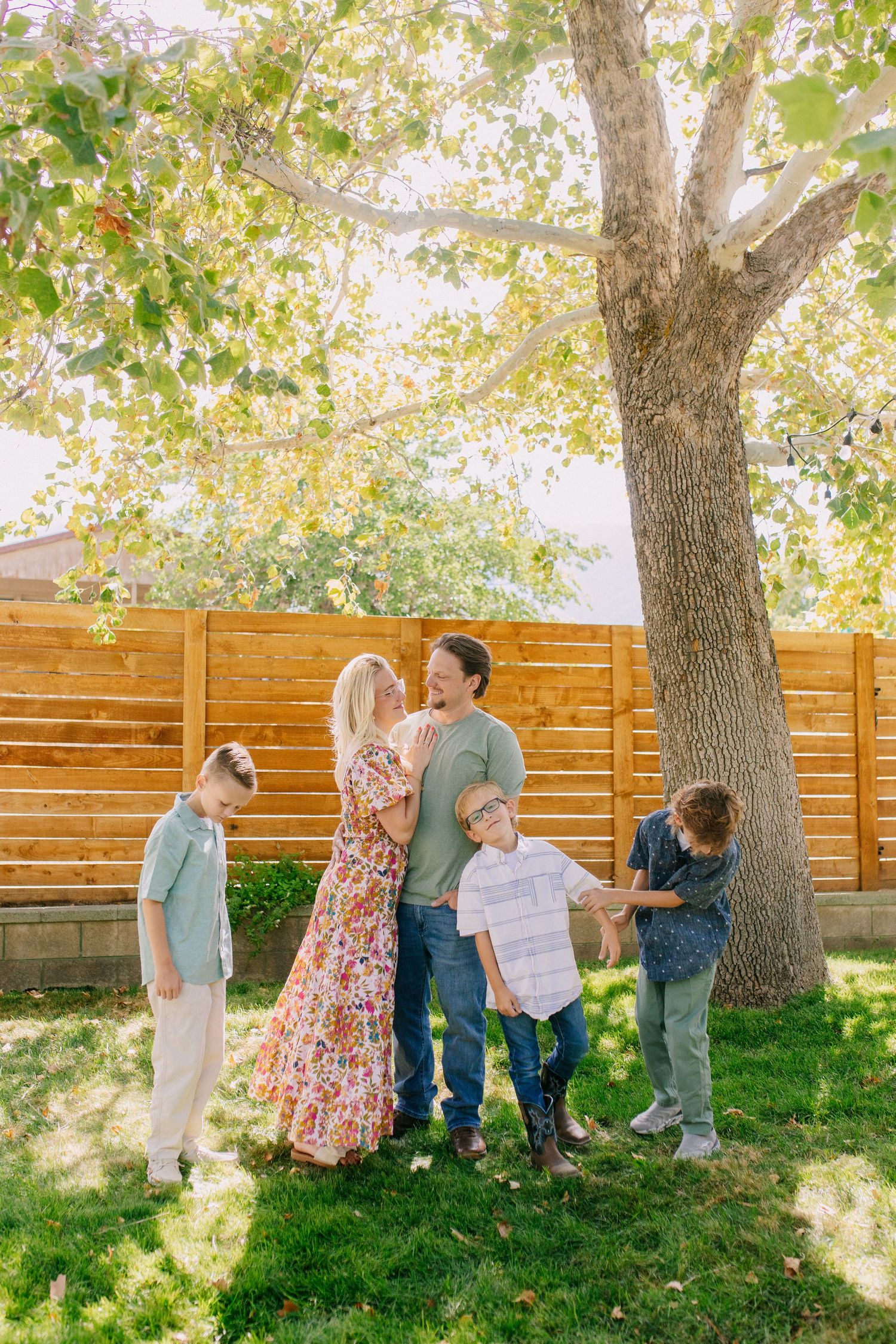 A sequence of candid outdoor family photos taken under large trees with a wooden fence backdrop in natural sunlight.