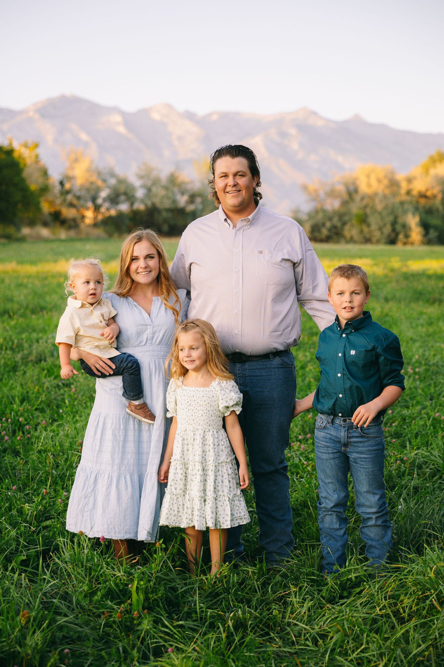 Family poses together in grassy field with mountains and sunset in background during outdoor portrait session.
