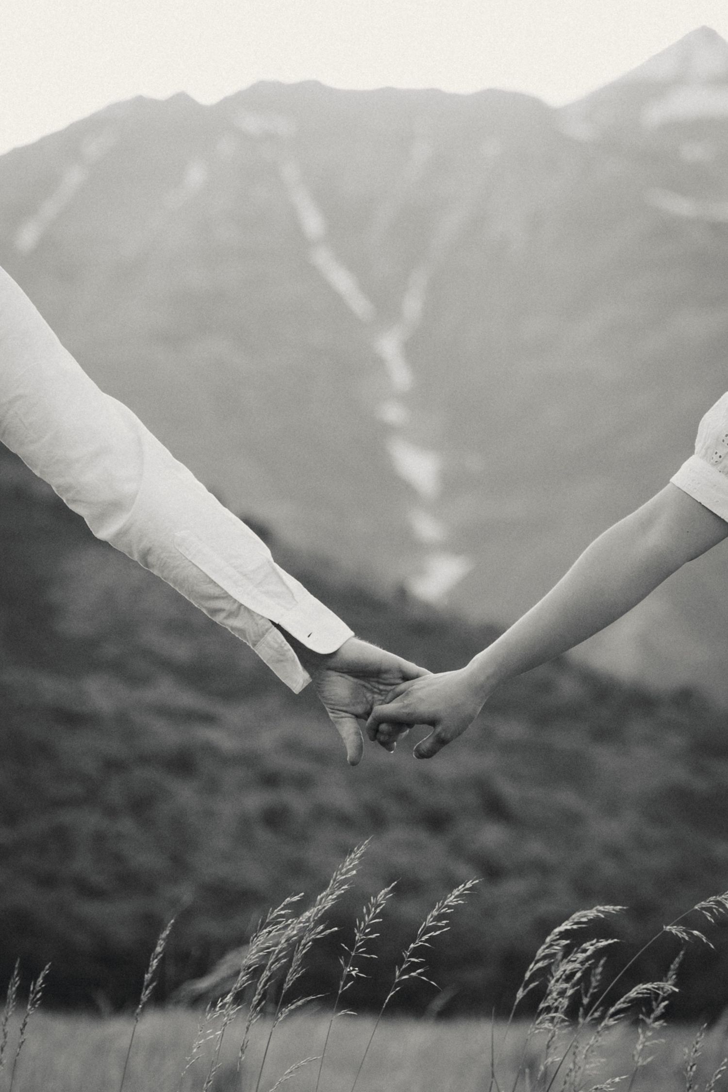Close-up of two hands holding each other with tall grass and mountains in the background.