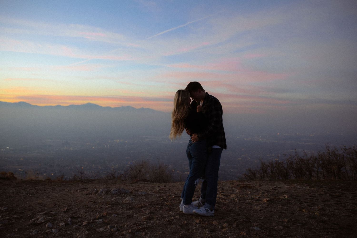 Silhouetted couple embracing against a colorful sunset sky over mountain landscape.