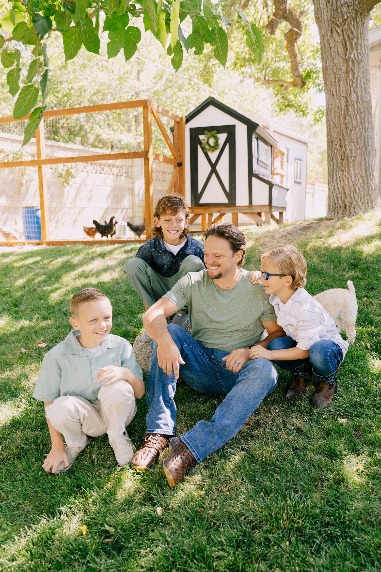 A group sits together on green grass under trees, sharing casual conversation and laughter on a sunny day.