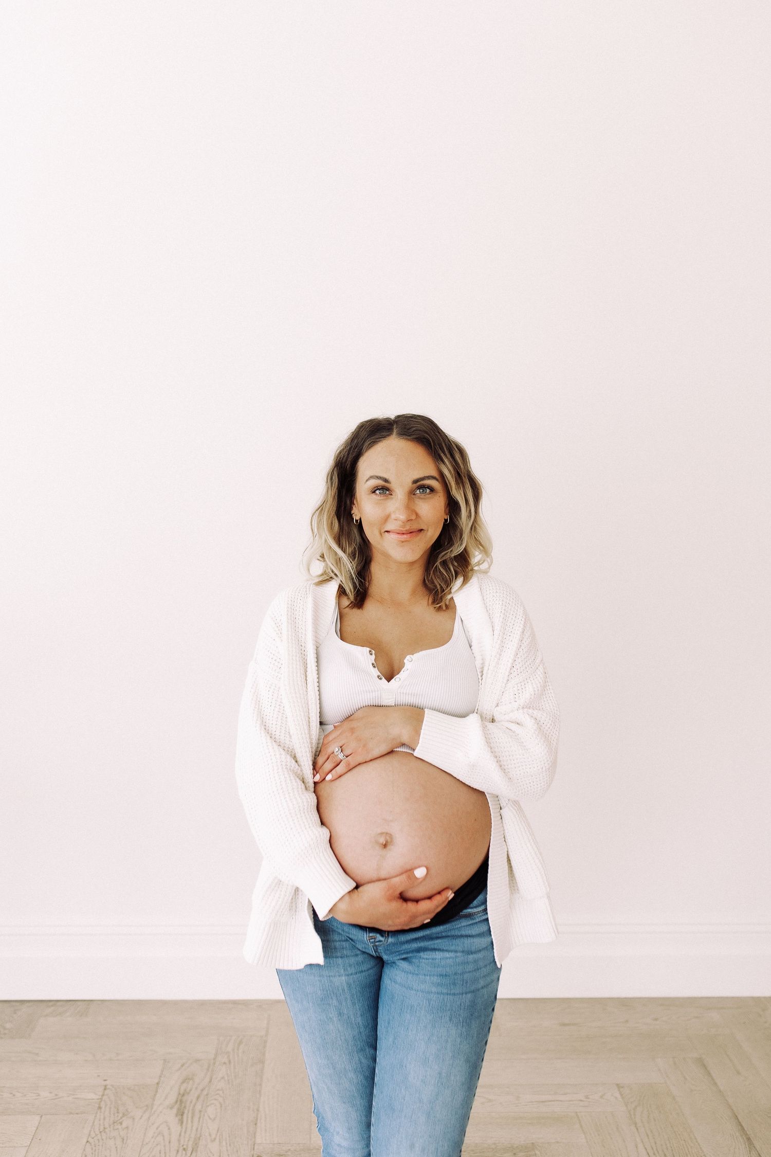 Maternity photoshoot showing a pregnant person in white shirt and jeans against a light background.