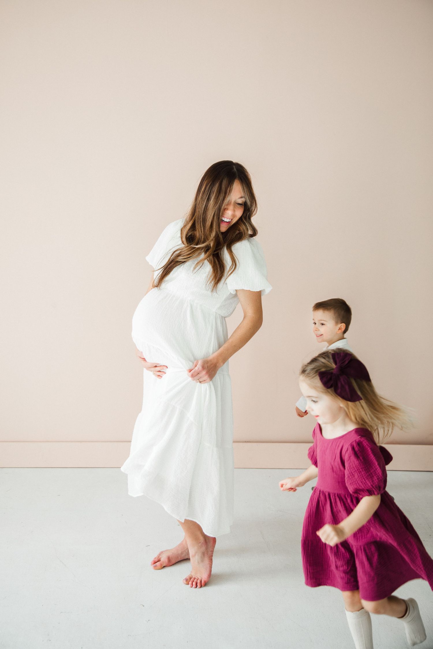 Children play around expectant mother in white dress during indoor photo session.