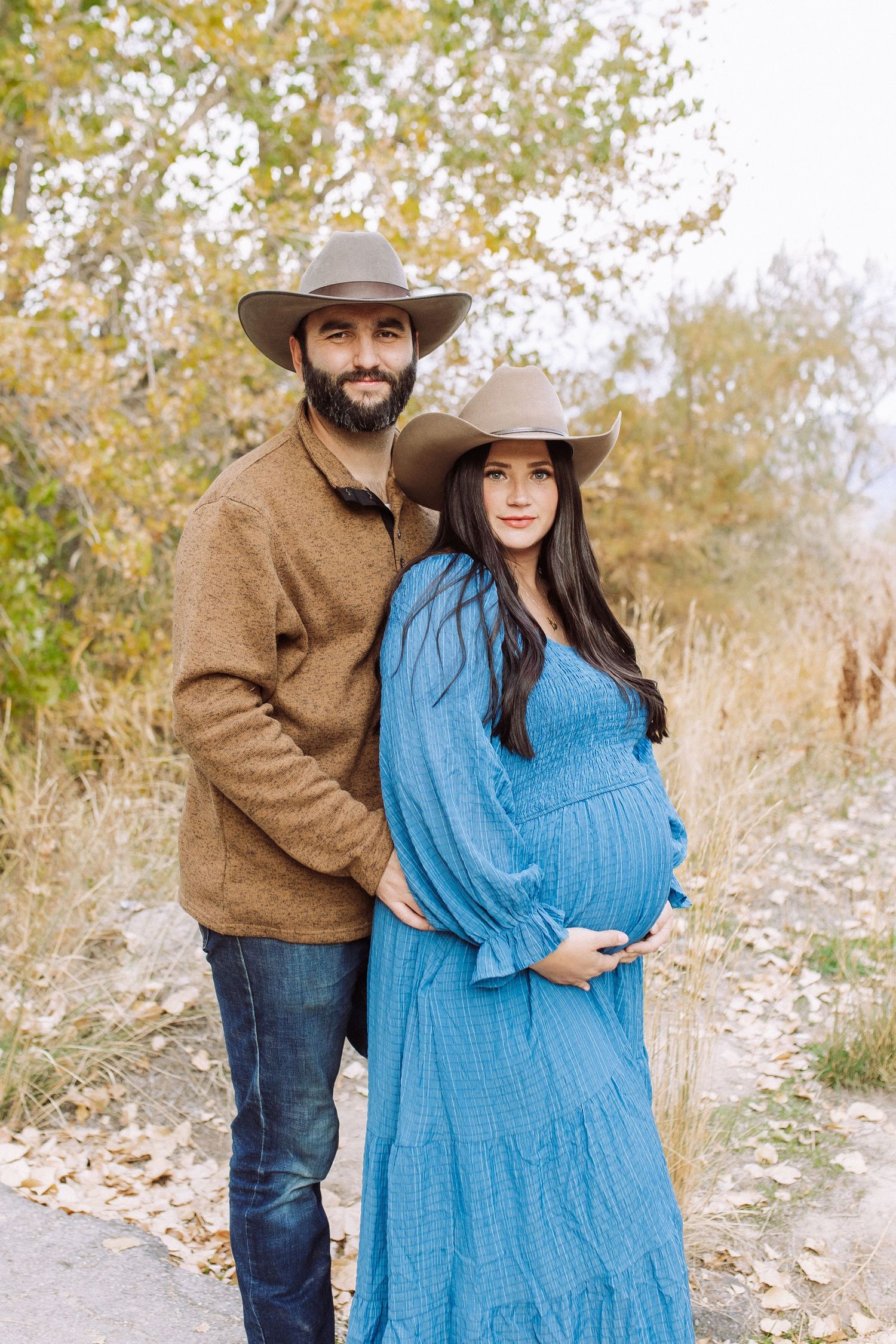 Couple in western wear poses for maternity photos on nature trail.