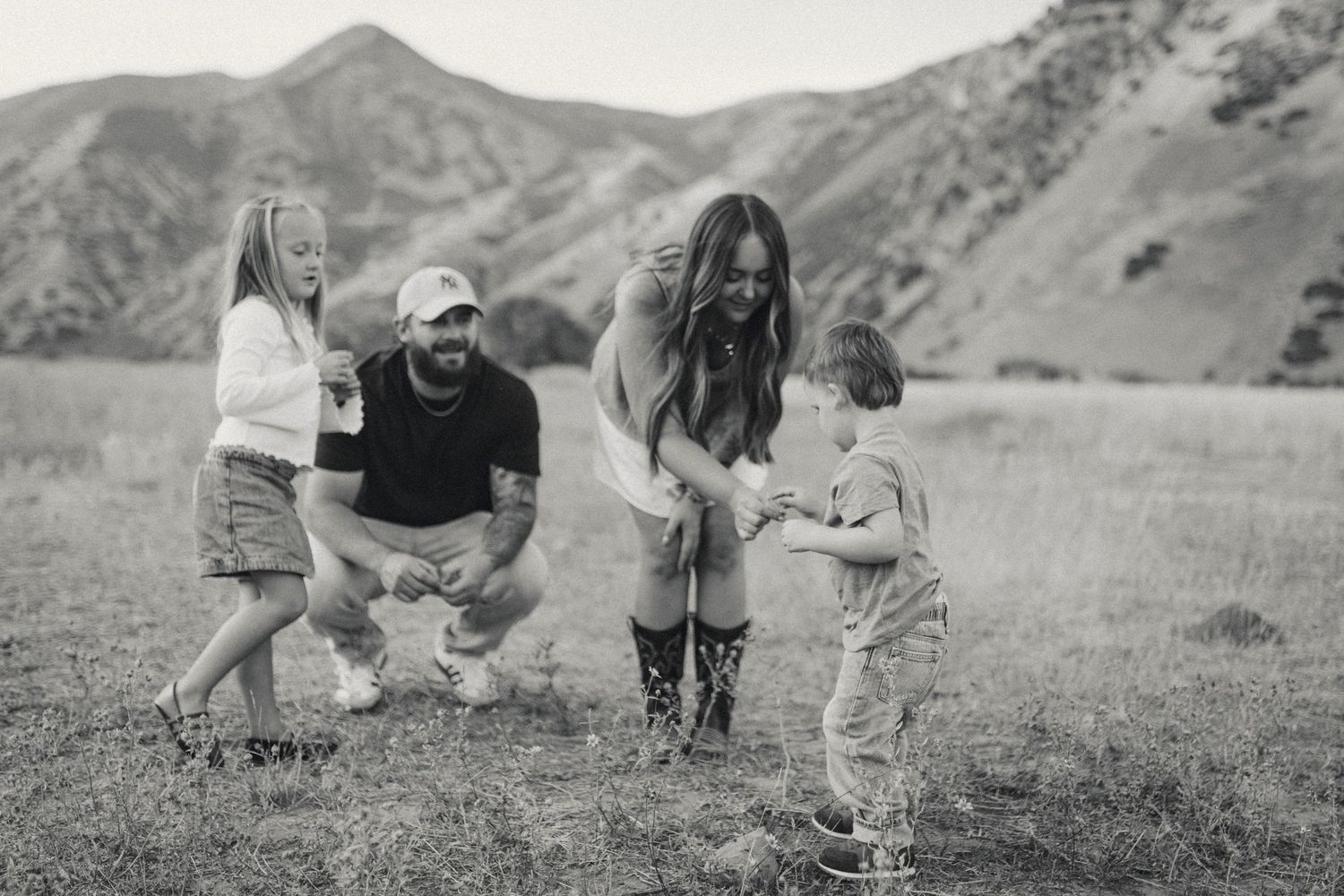 A group of people crouch down to examine something interesting in a grassy mountain landscape.