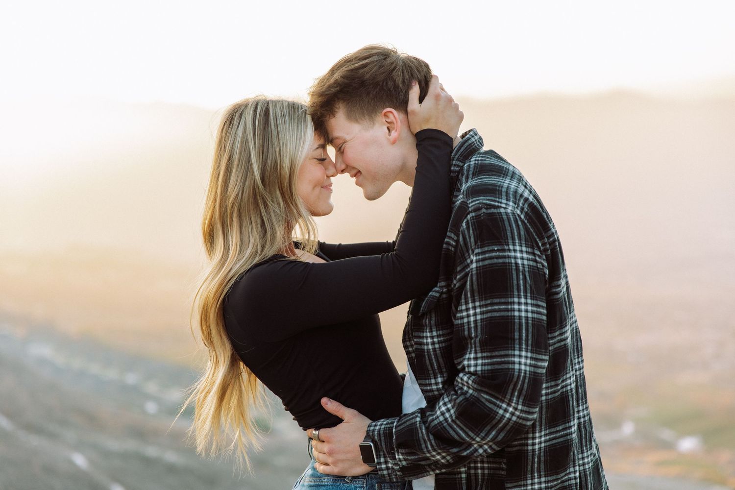 A couple embraces on a hilltop during golden hour wearing casual clothing.