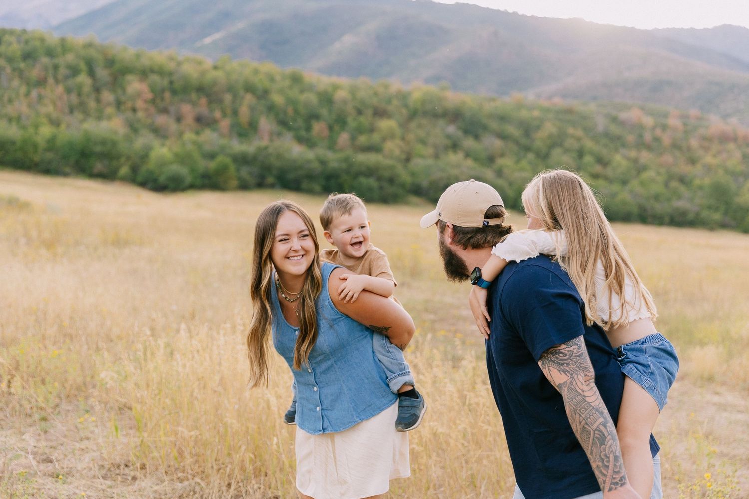 A group of people embrace and laugh together in a golden field with mountains in the background at sunset.