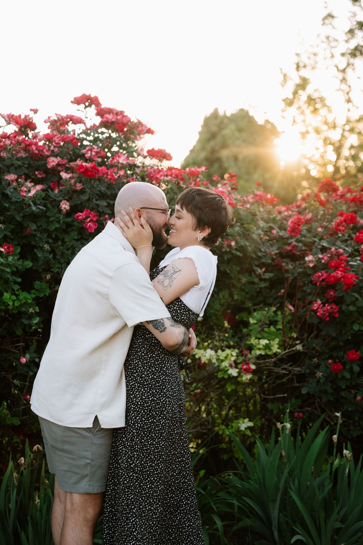 A romantic couple shares an intimate moment in a garden with blooming red flowers and sunset lighting.