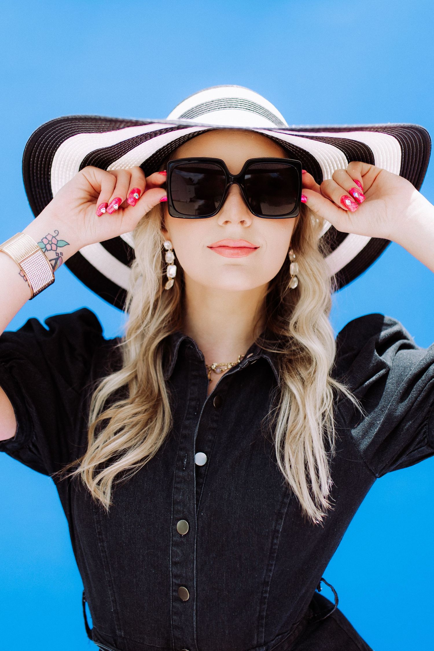 Stylish woman in black denim dress and striped sunhat poses against bright blue background.
