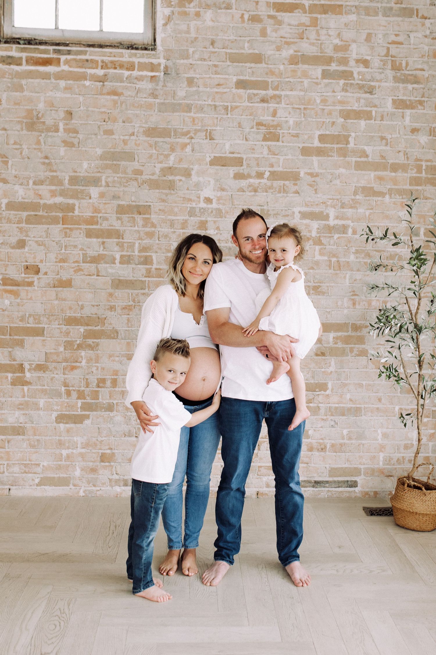 Family maternity photoshoot in front of exposed brick wall wearing white shirts and jeans.