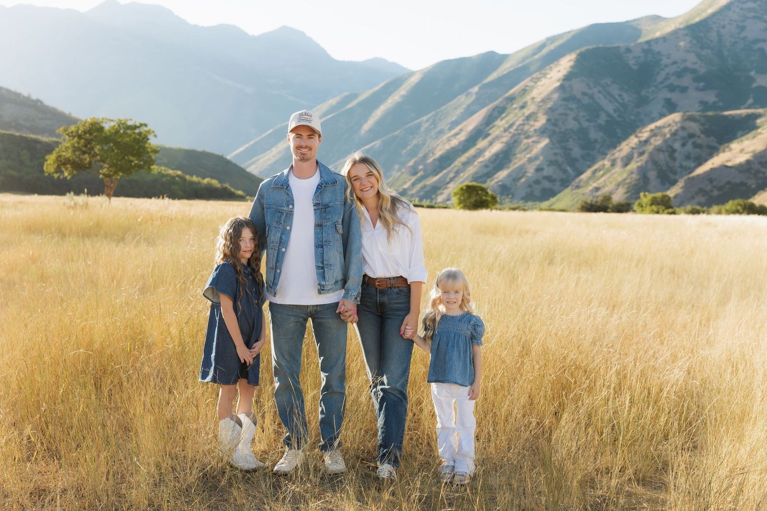 Family in coordinated blue and white outfits walking through golden wheat field at sunset with mountains in background.