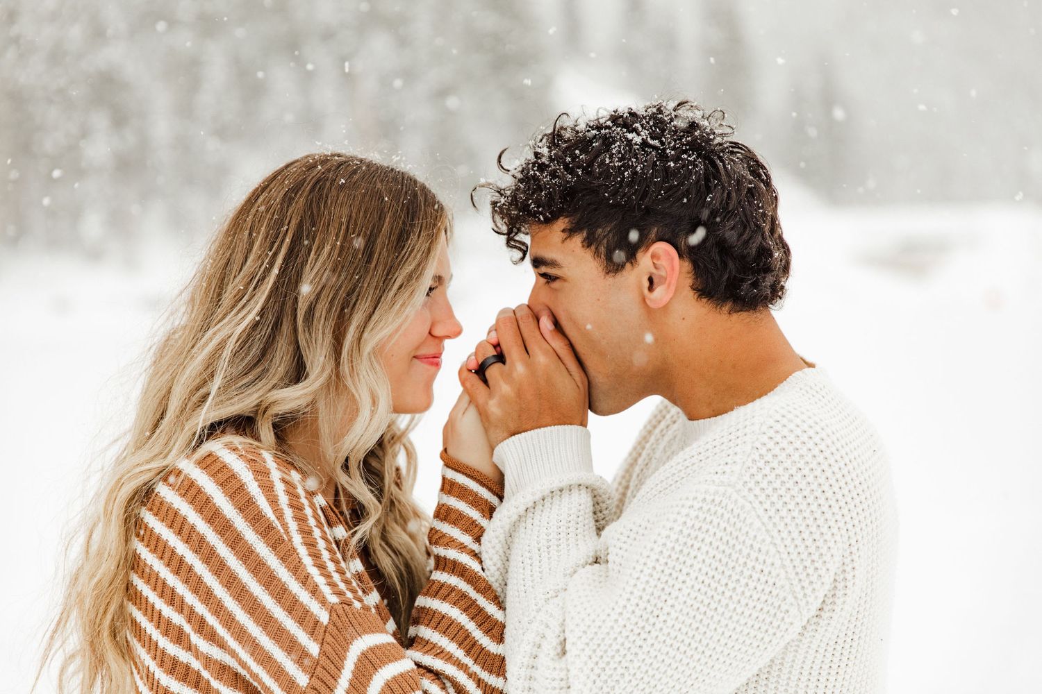 A couple in winter clothing shares a tender moment against a snowy forest background.