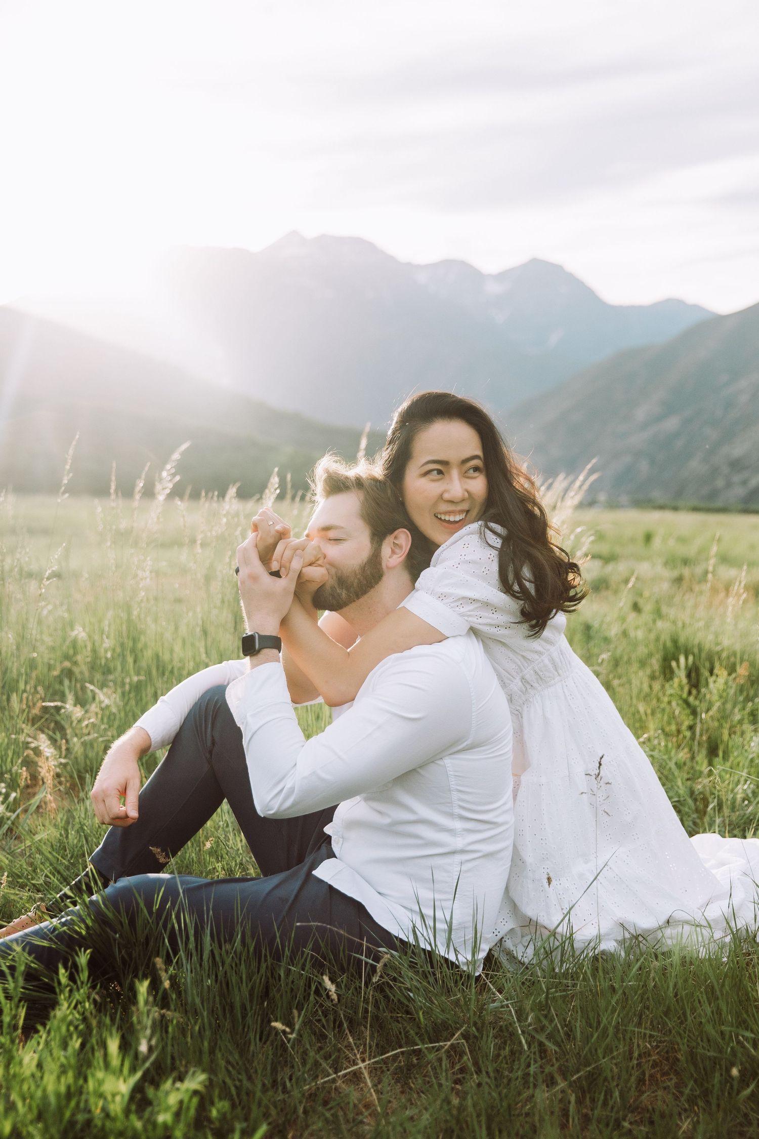Couple lying in green meadow with mountain backdrop during sunset photo session.