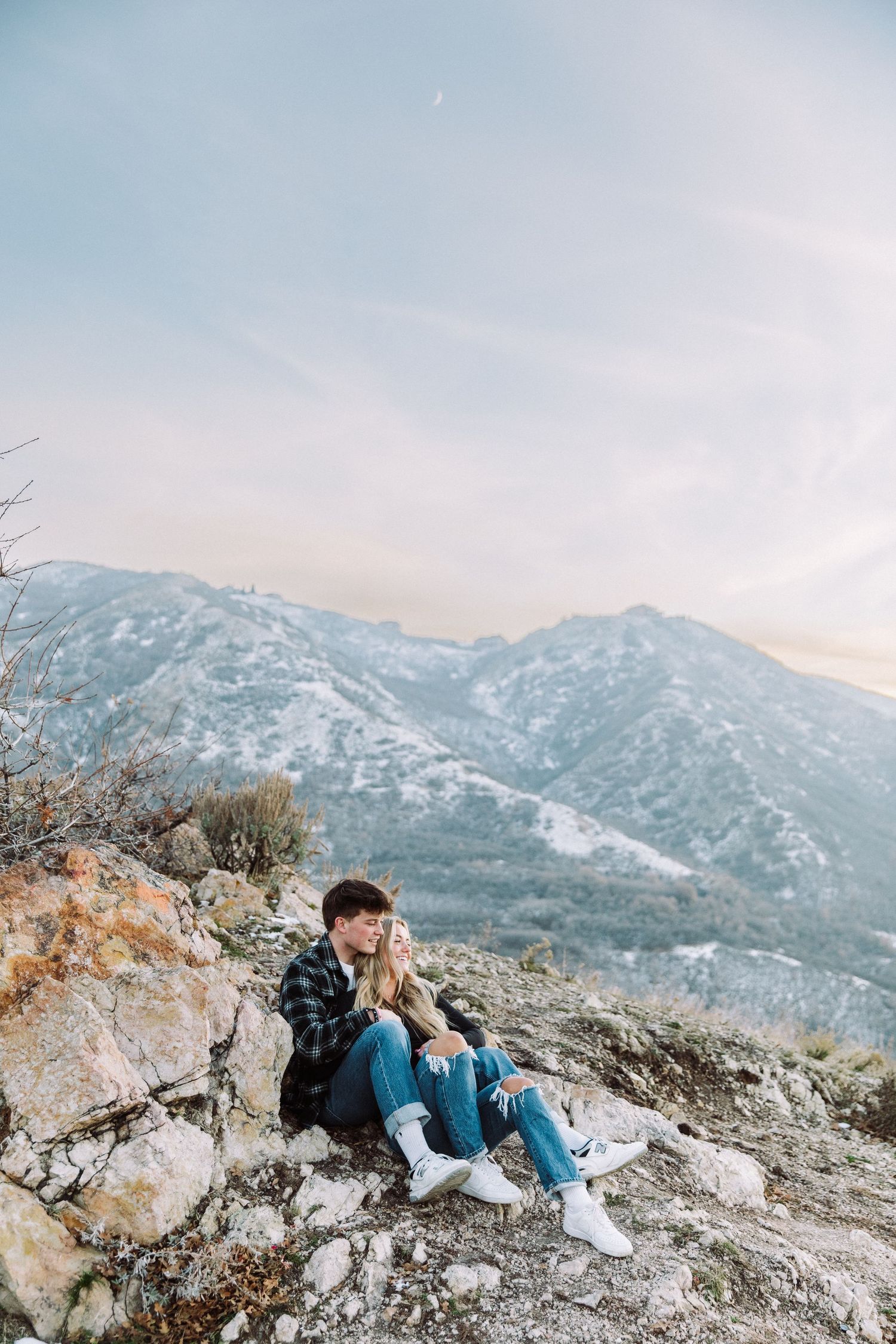 Couple relaxing on rocky mountain ledge with snow-capped peaks in the distance.