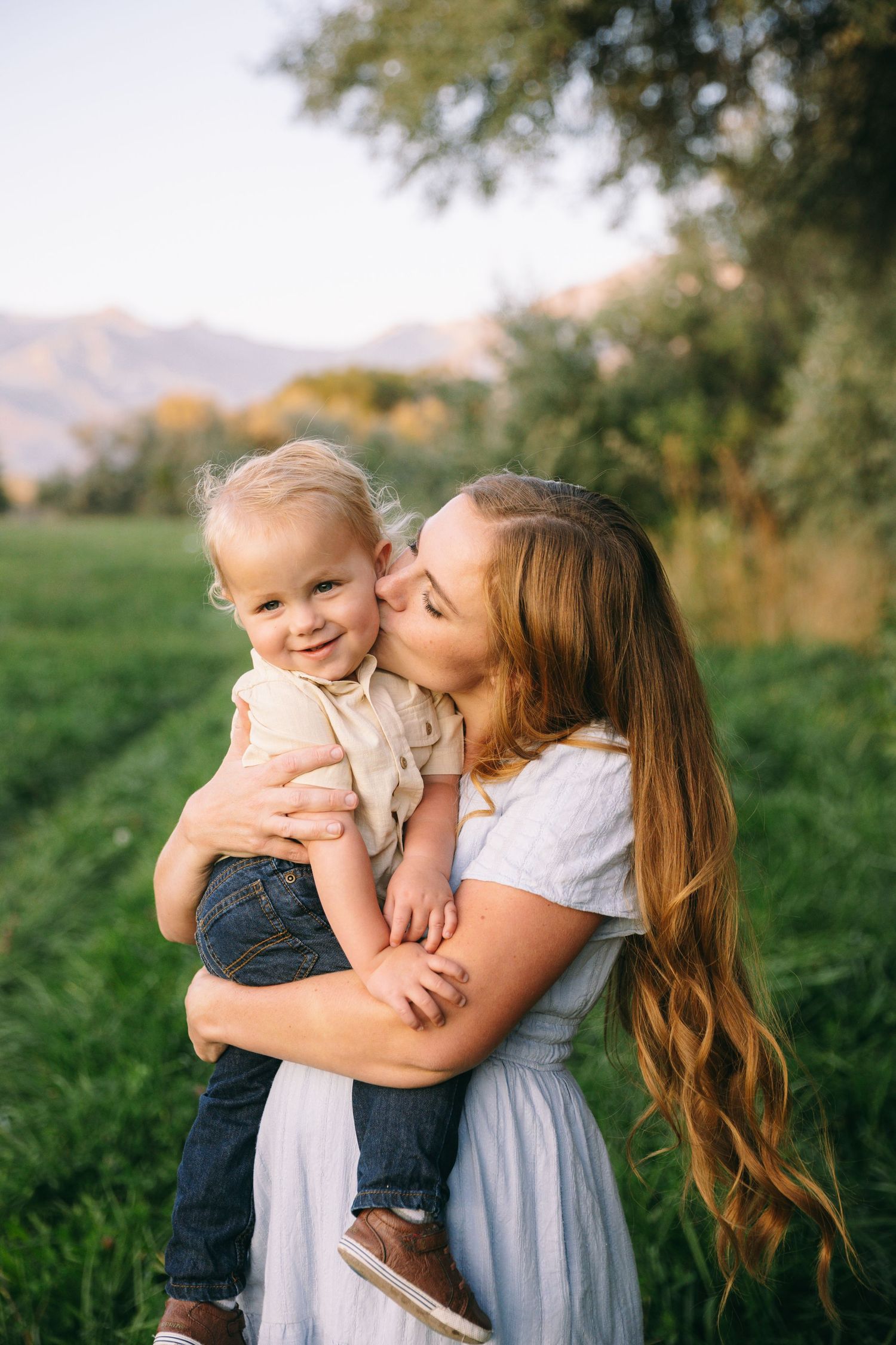 A tender moment between parent and child outdoors in a grassy field with mountains in the background.