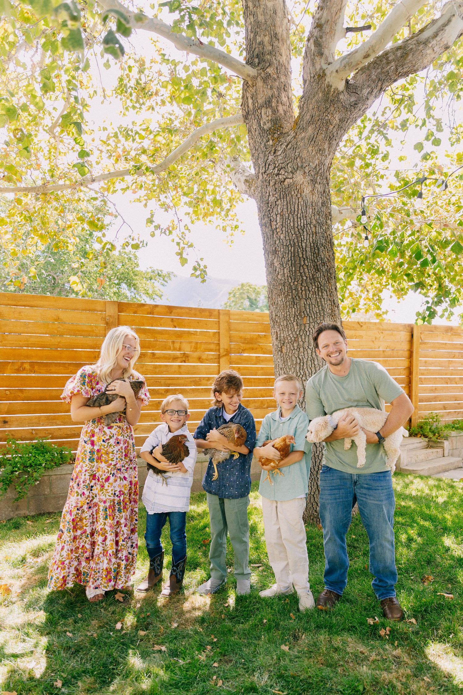 A group plays together under autumn trees in front of a wooden fence during an outdoor family photoshoot.