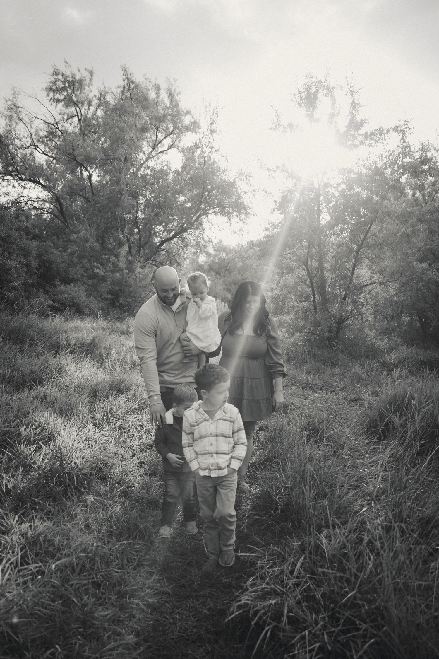 Black and white portrait of figures walking through tall grass with sunlight filtering through trees.