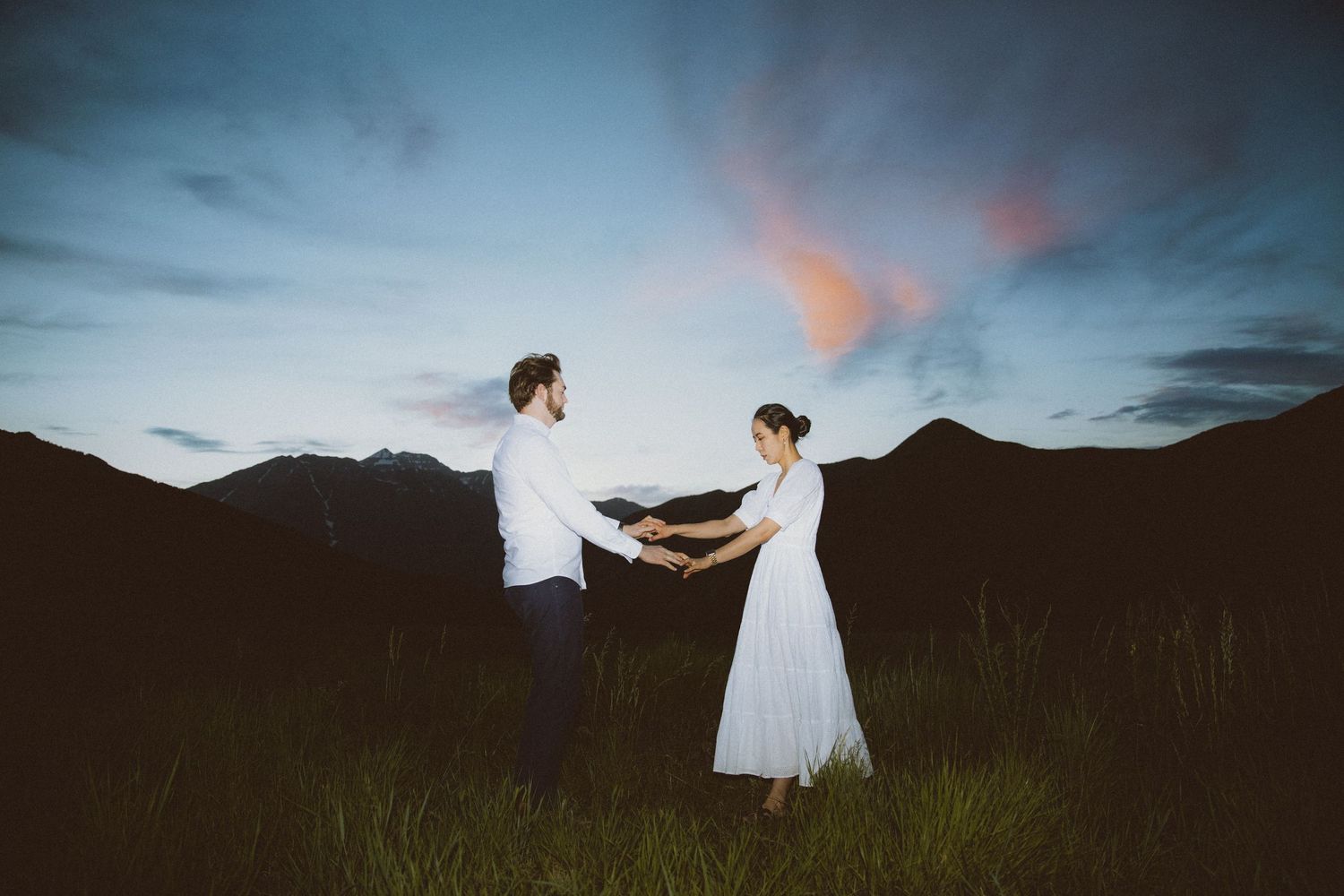 Two people in white clothing dance in a field at sunset with mountains in the background.