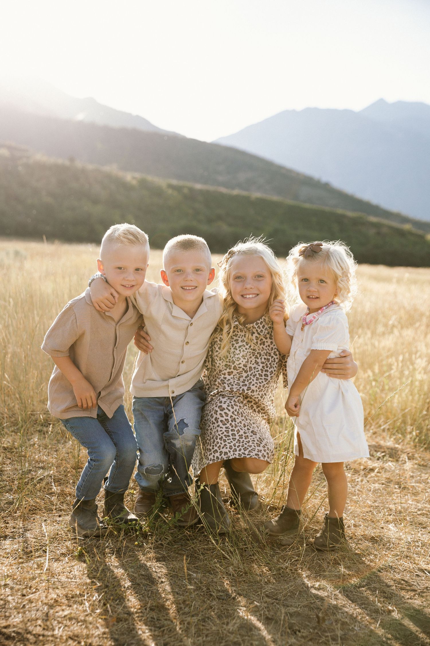 Four small children sit together laughing in a golden wheat field at sunset with mountains in background.