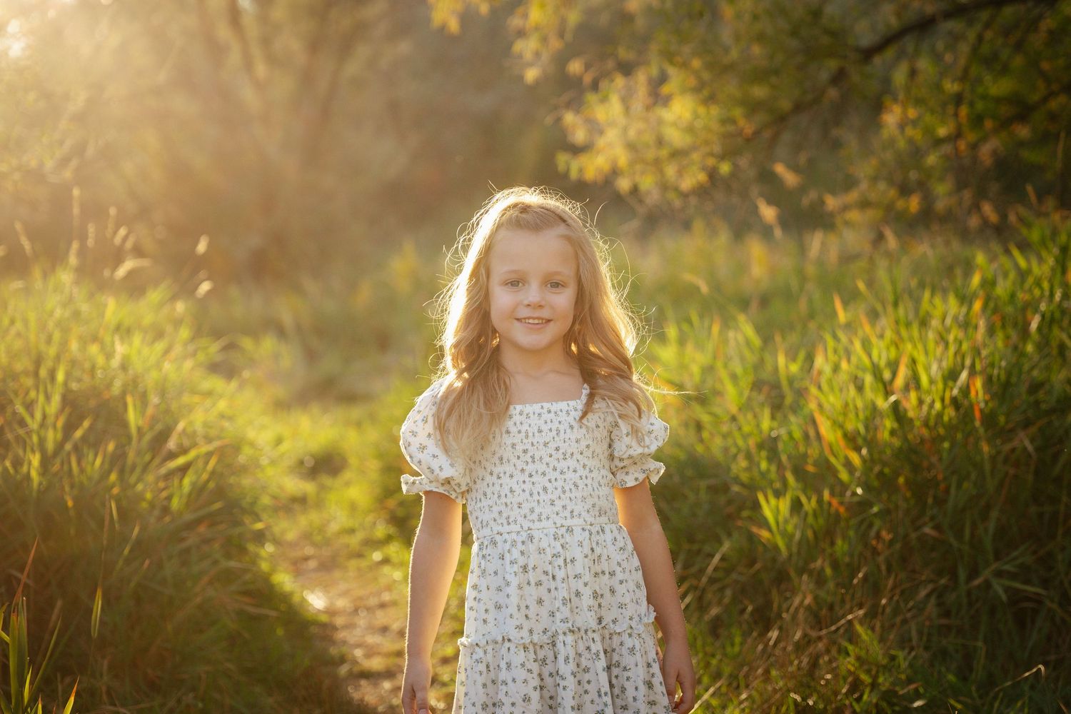 A person stands in a sunlit meadow during golden hour wearing a floral dress.