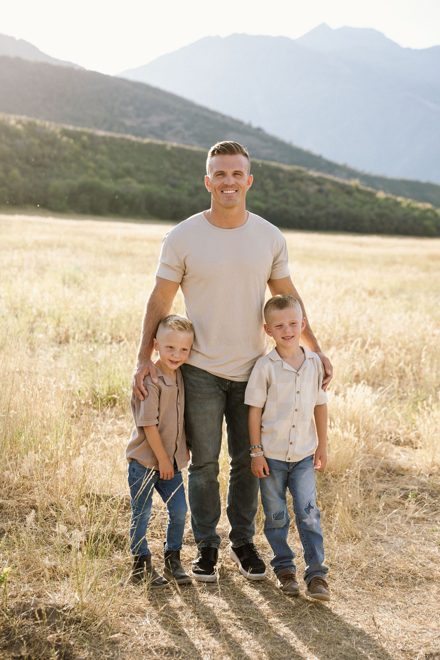 Three figures stand together in a golden field with mountains in the background during sunset.