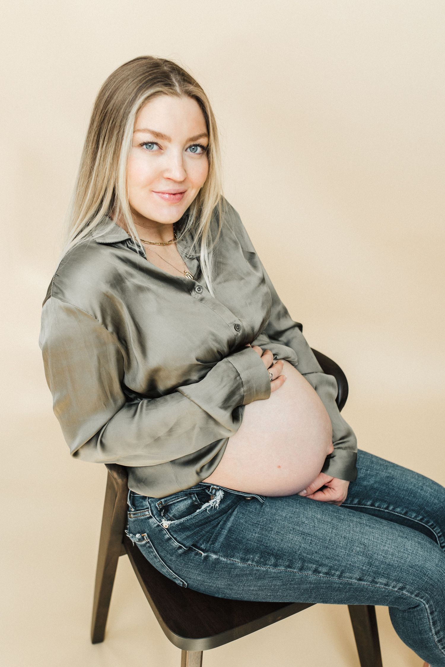 Pregnant woman in silk shirt and jeans sits on wooden chair during photo session.
