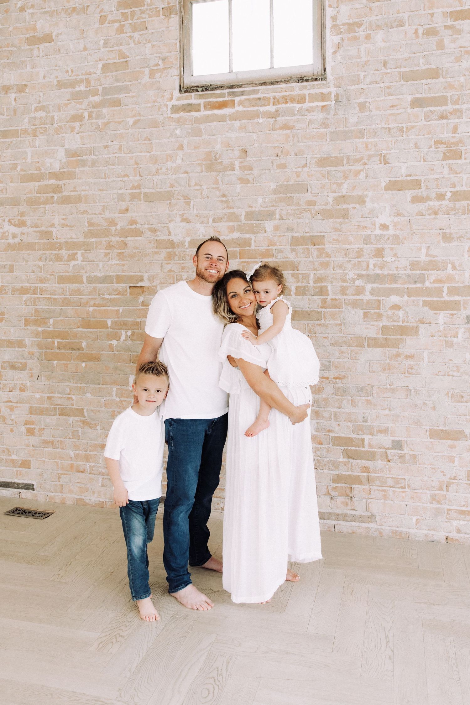 Family in white clothing poses together against exposed brick wall in minimalist setting.