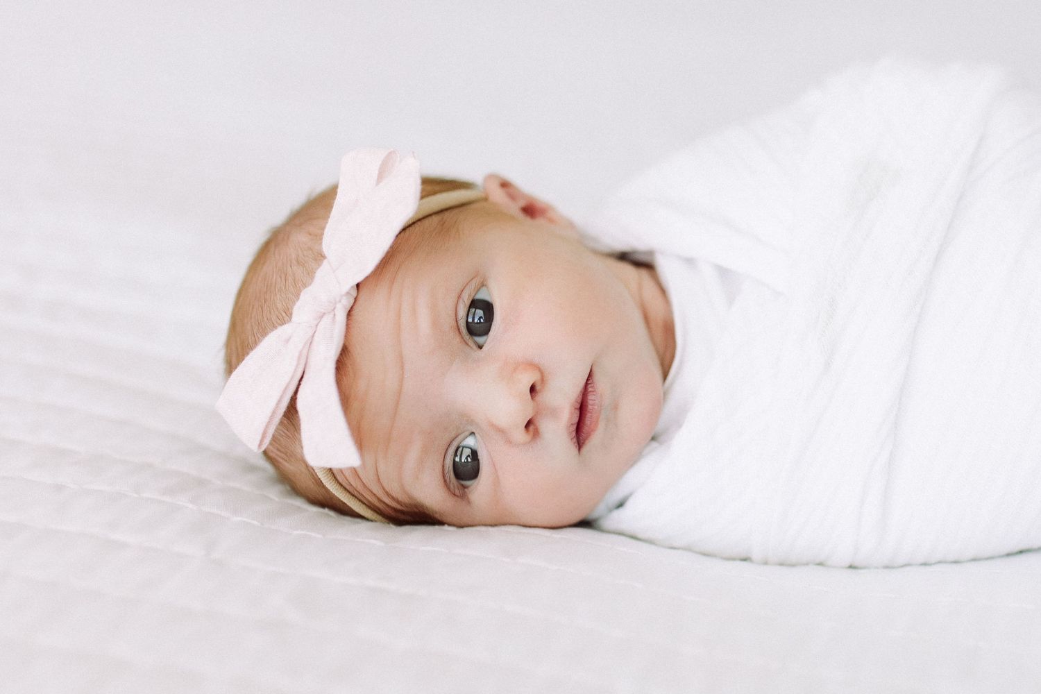 Close-up portrait of an infant wearing a pink bow headband against white bedding.