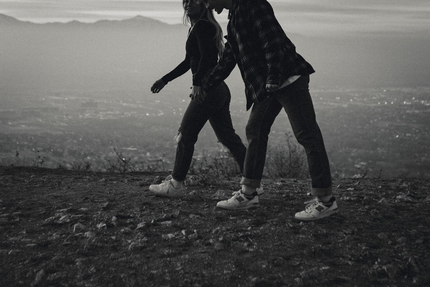 Black and white photo of two people's legs walking together on hilltop at dusk.