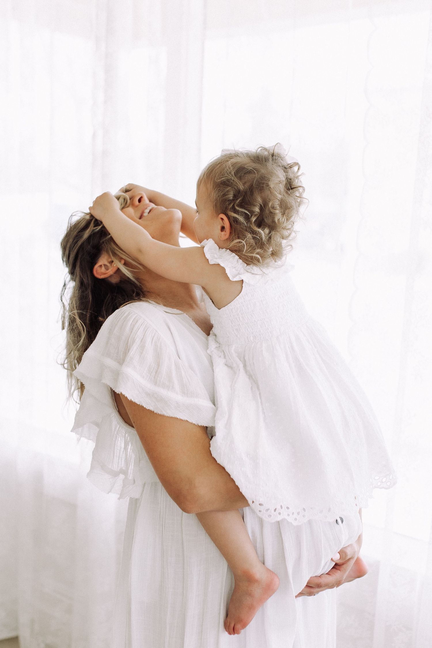 Mother in white dress lifts joyful child in front of bright window.