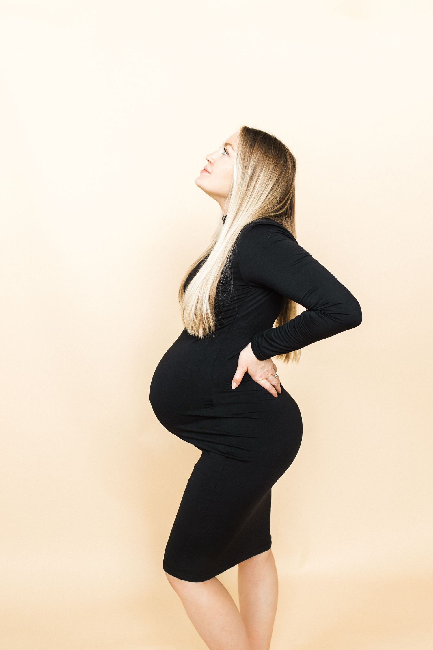 Side view of pregnant woman in black dress against cream backdrop during studio shoot.