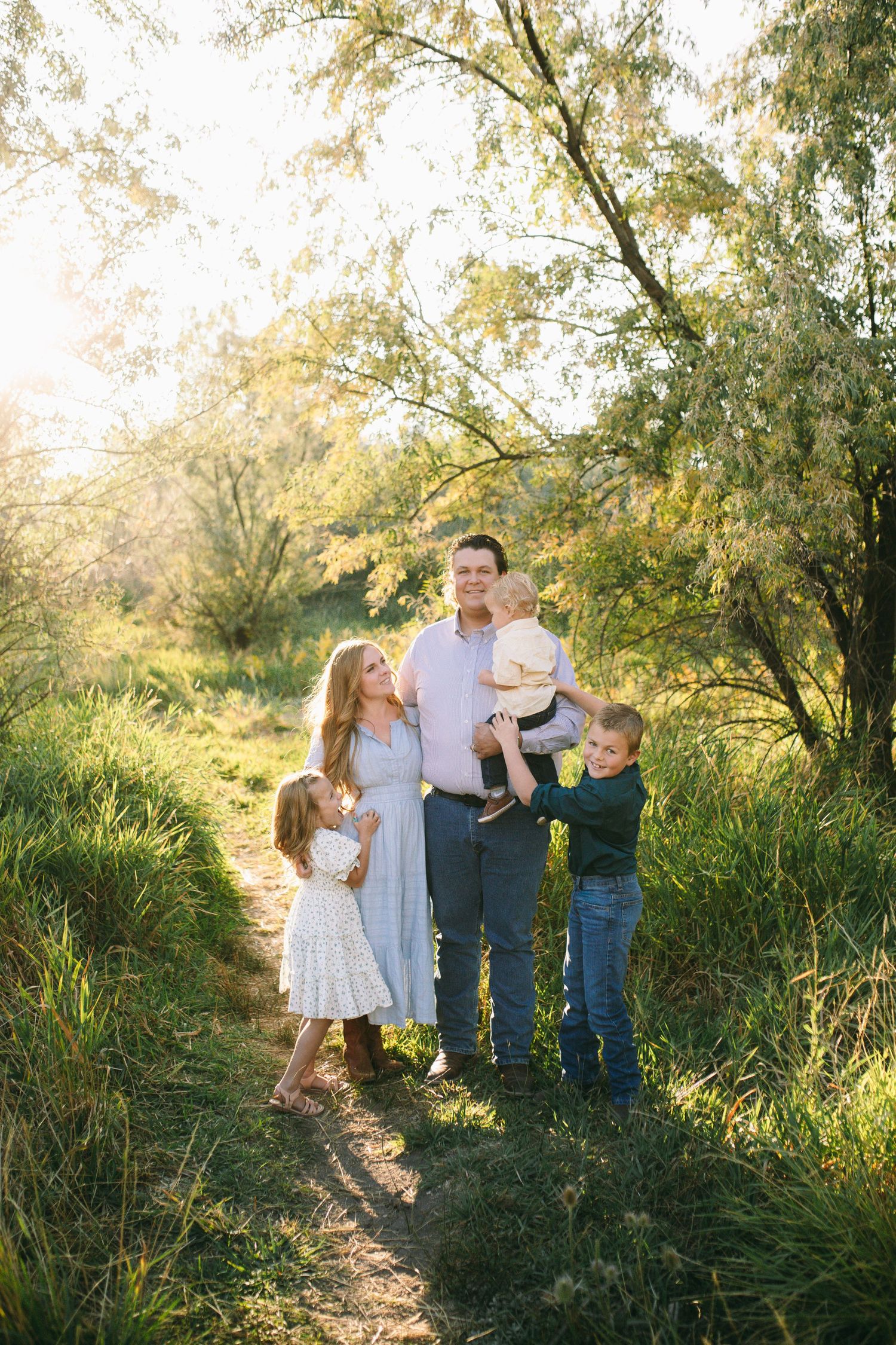 A family in coordinating blue and white outfits poses together on a sunlit nature path surrounded by tall grass and trees.