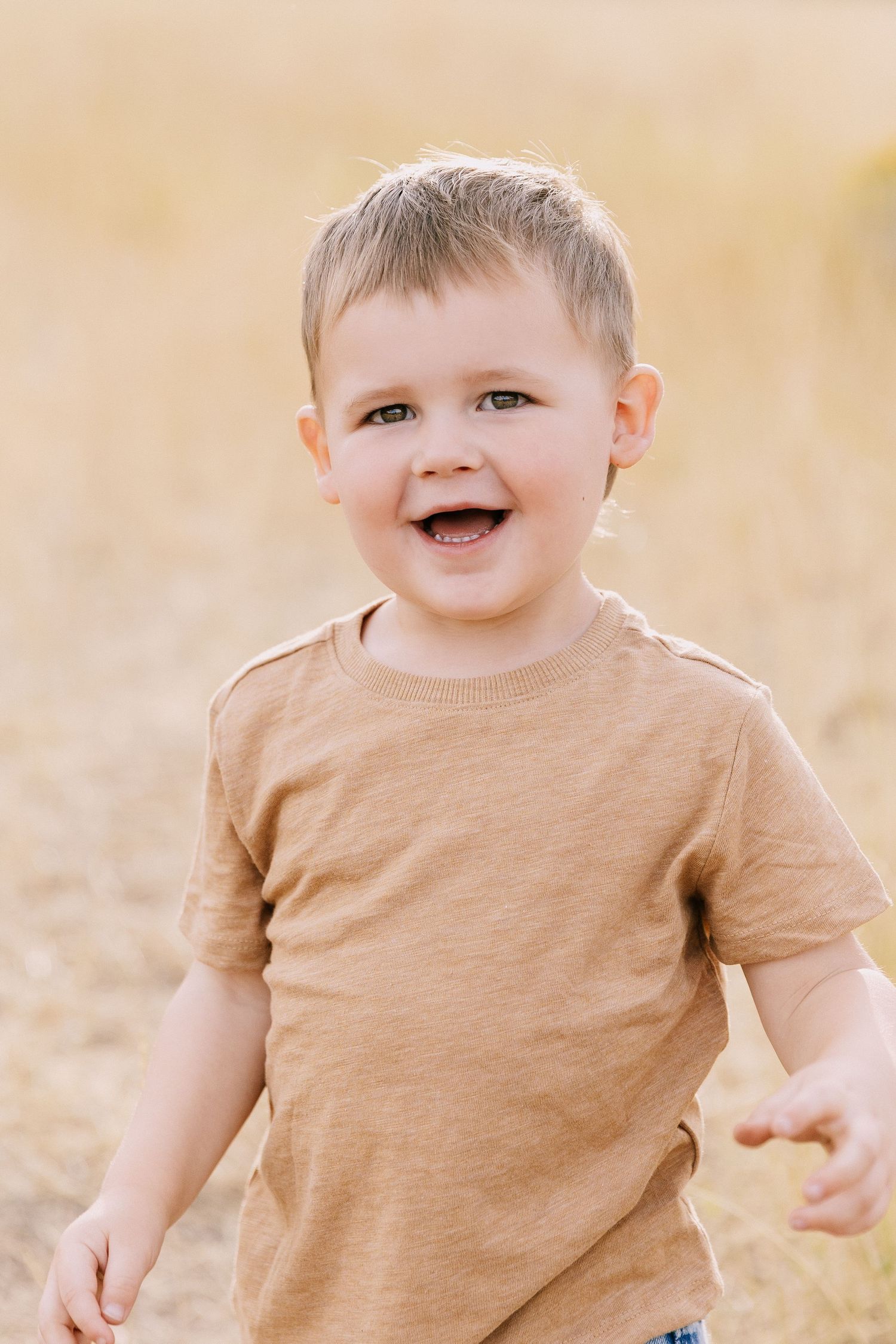 A young child wearing a tan t-shirt plays outdoors in warm sunlight with a joyful expression.