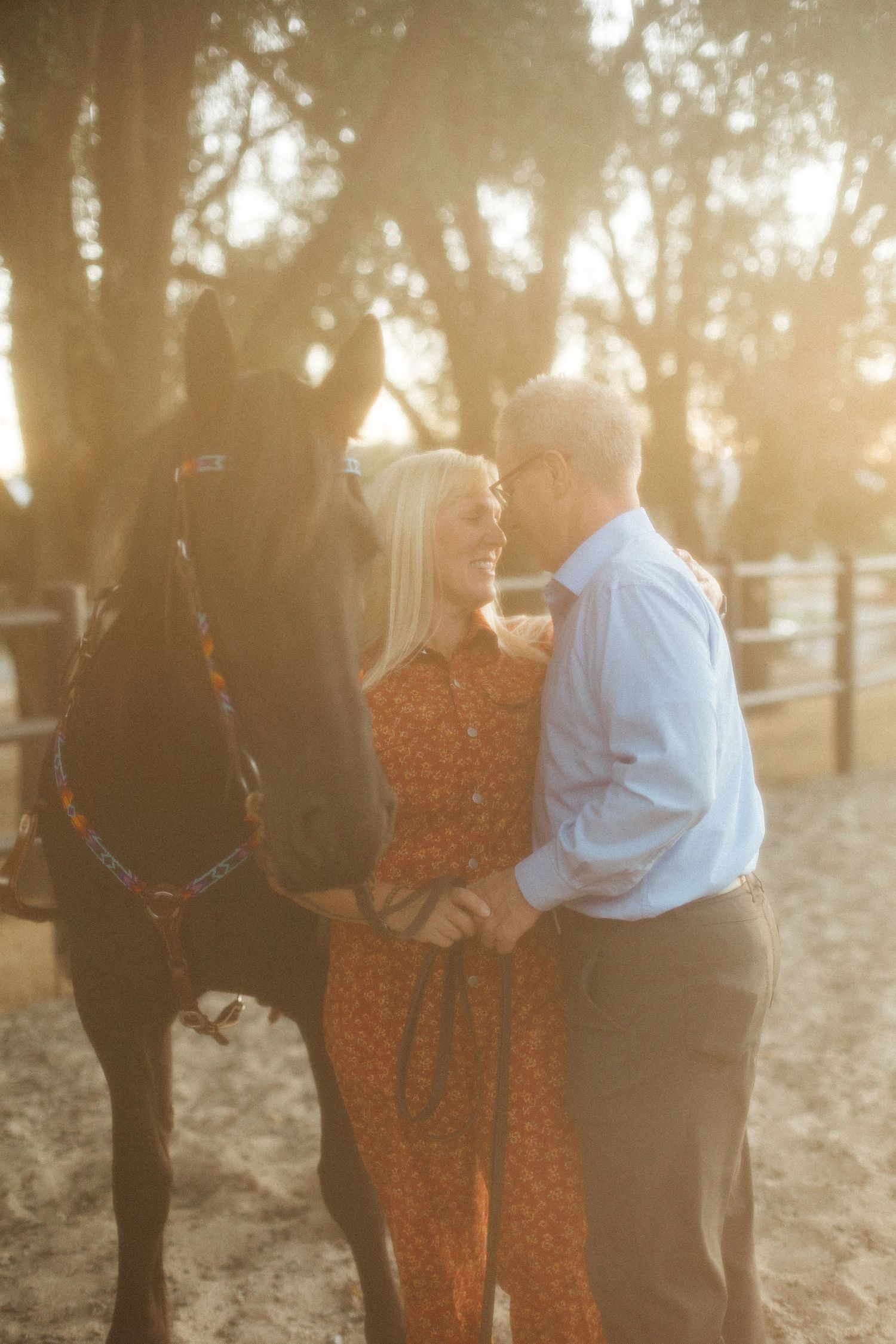 A couple stands close together next to their black horse during a golden sunset.