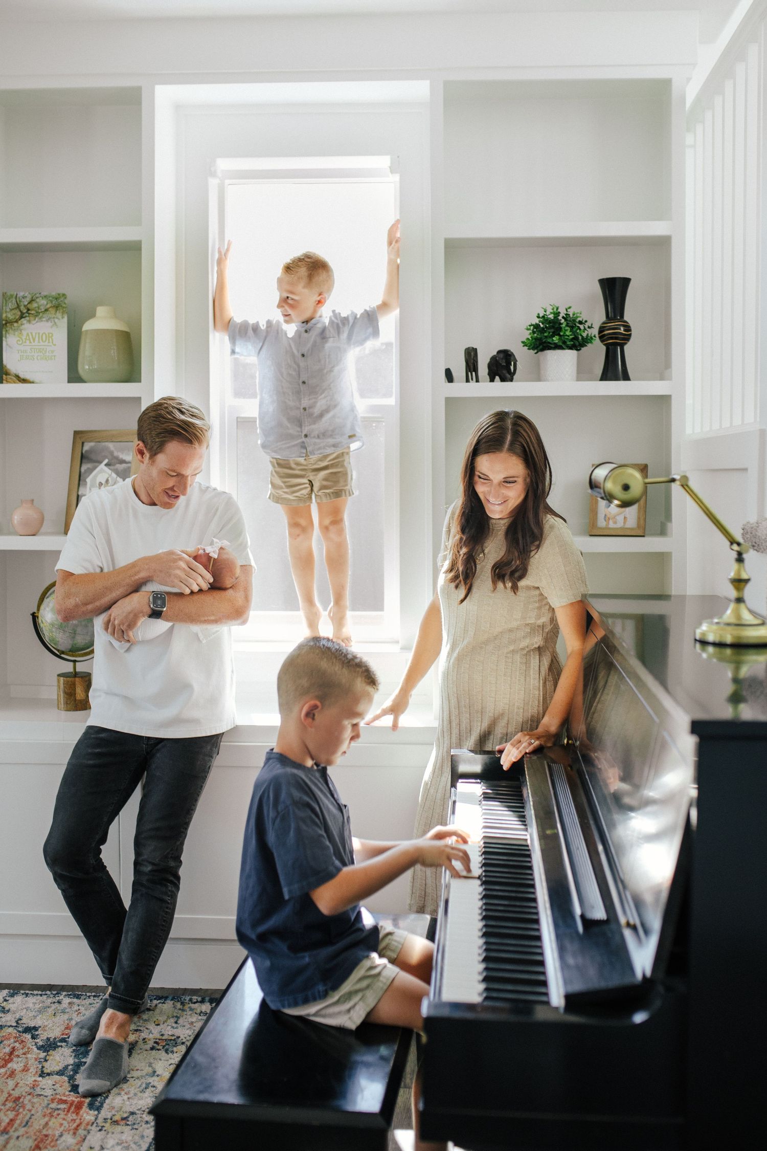 Family playing and dancing together around a piano in a bright white room with built-in shelving.