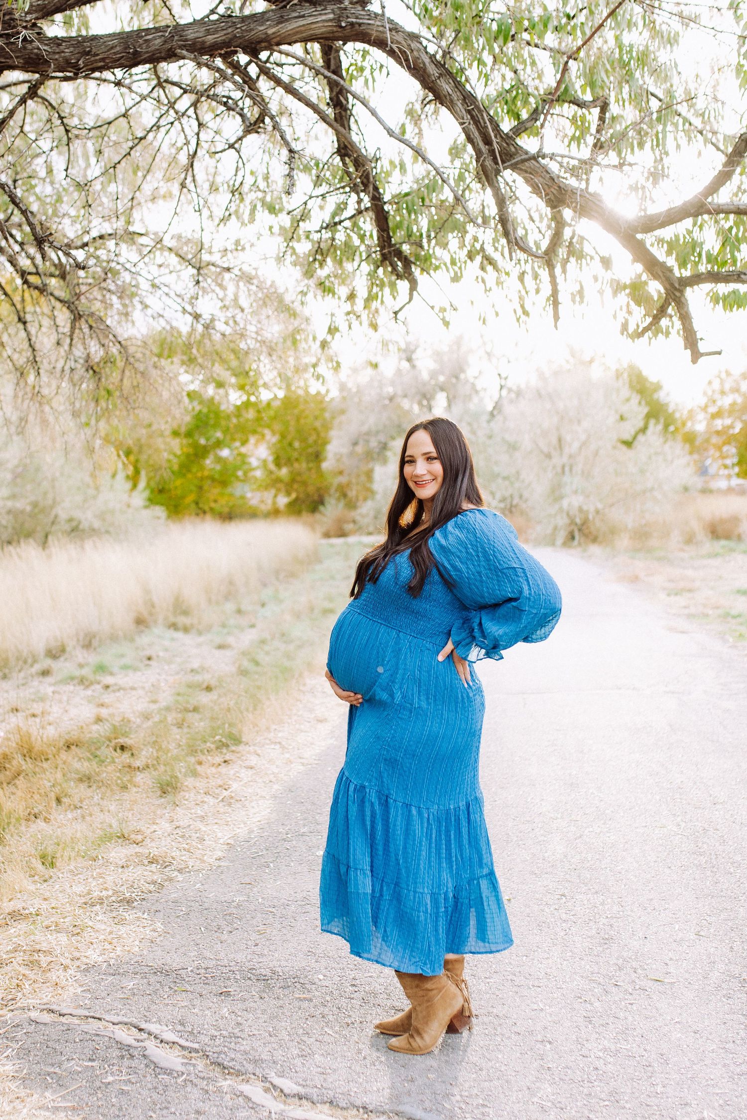 Woman in flowing blue dress twirls on dirt path during outdoor maternity photoshoot.