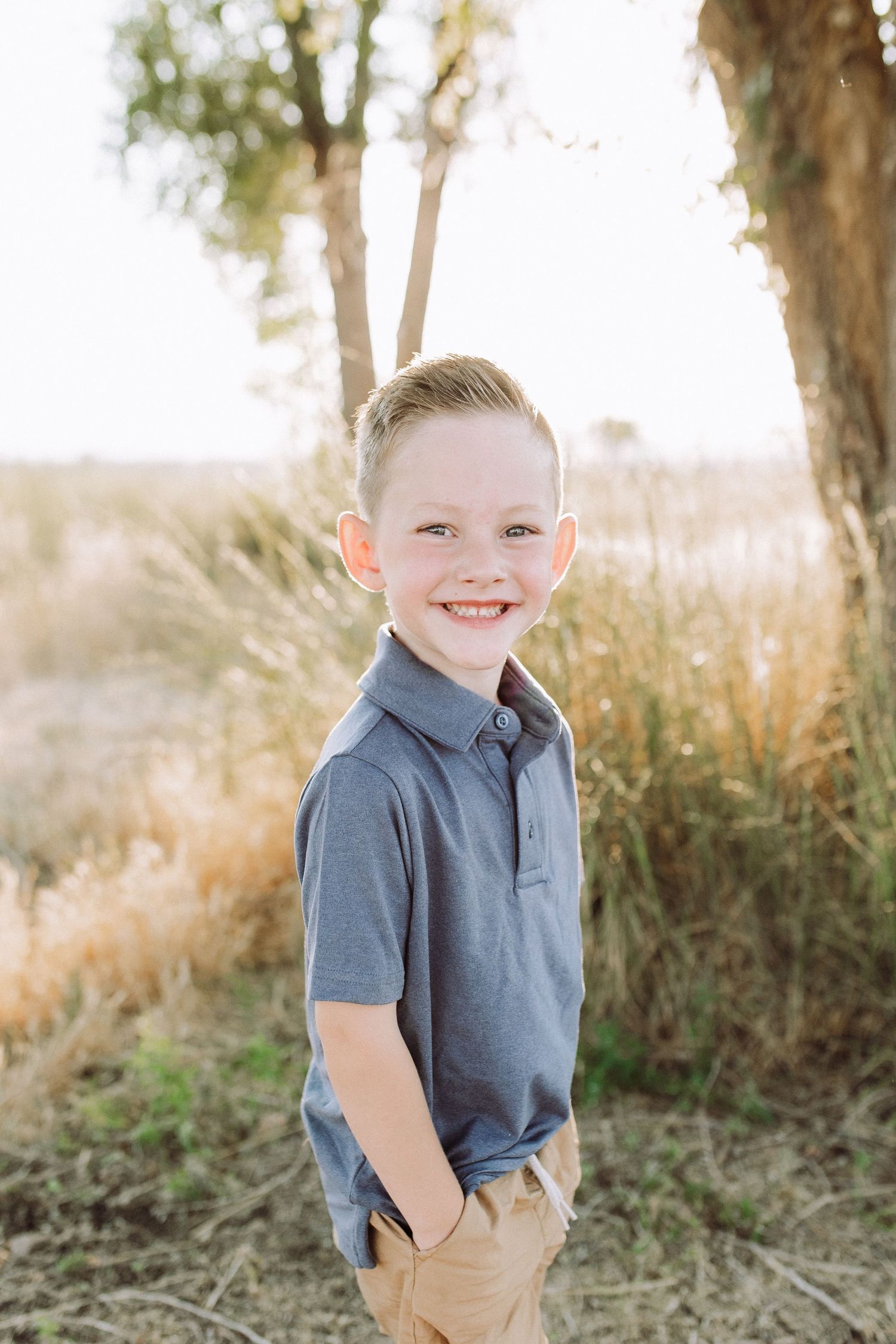 Young boy in a grey shirt poses outdoors with a bright smile against a natural backdrop.