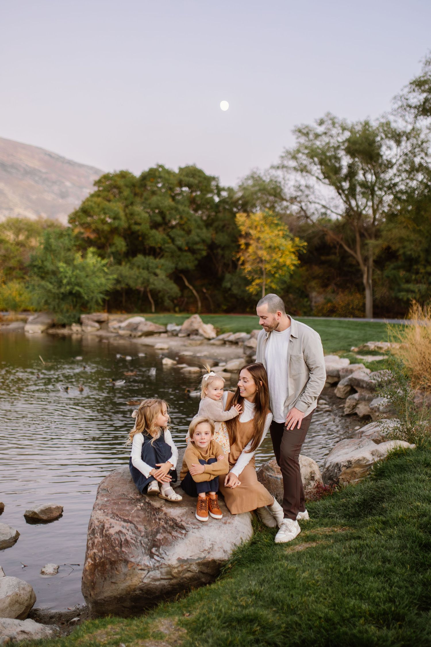 Family gathered on rocks by a lake during sunset with mountains in background.