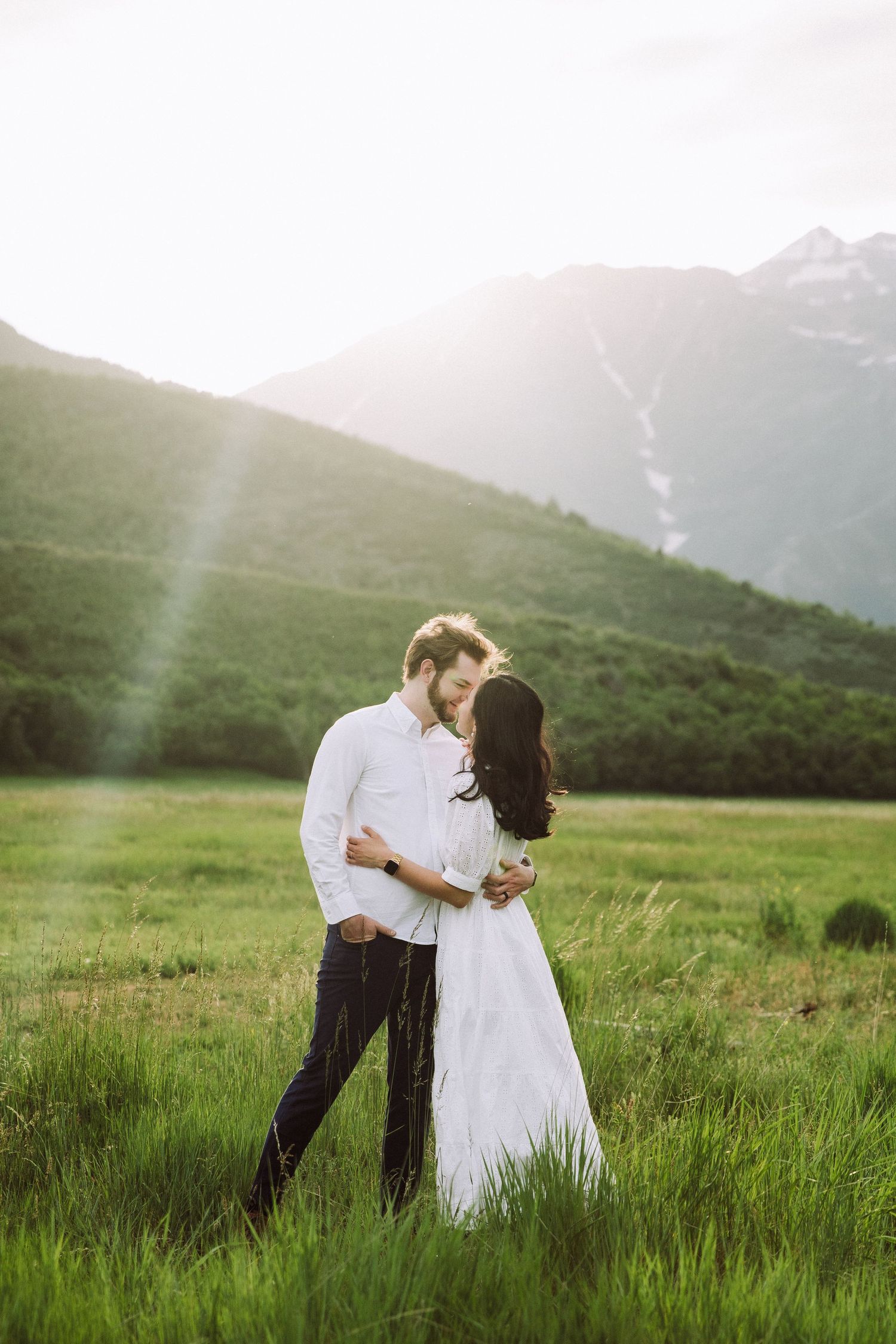 Romantic mountain engagement photo with couple embracing in sunlit meadow.