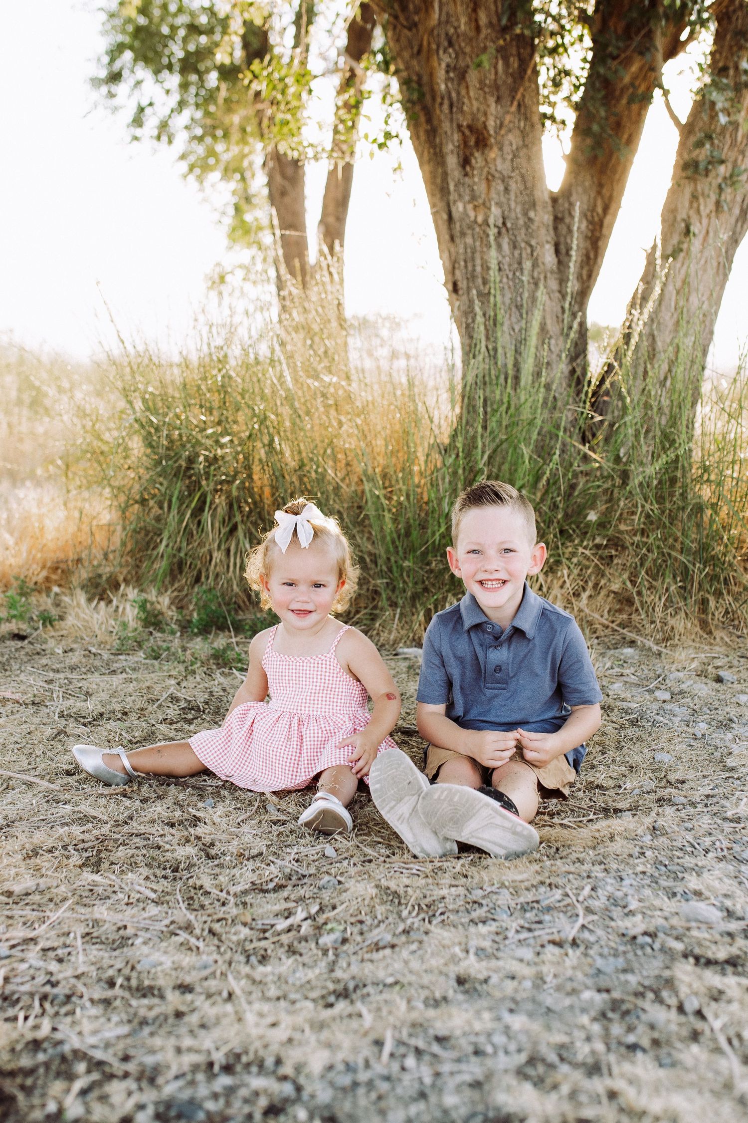 Two children sit together under tall trees on a sunny day in a natural outdoor setting.